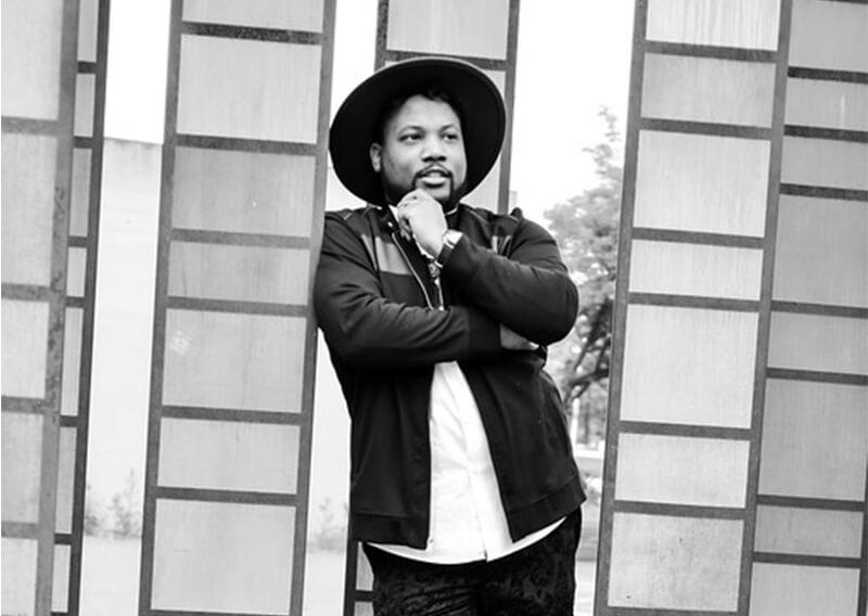 Jimmy Shields in a black and white photo with black fedora hat leaning on a post. 